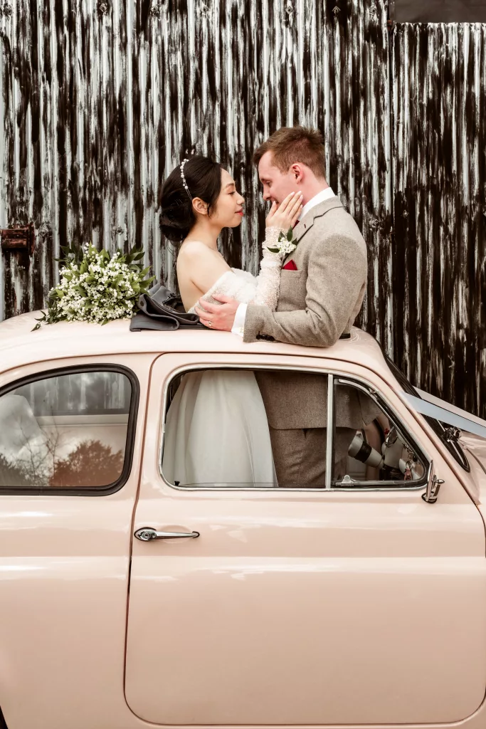 Bride and groom in a pink Fiat 500 standing in front of the black Cripps Barn outer wall.
