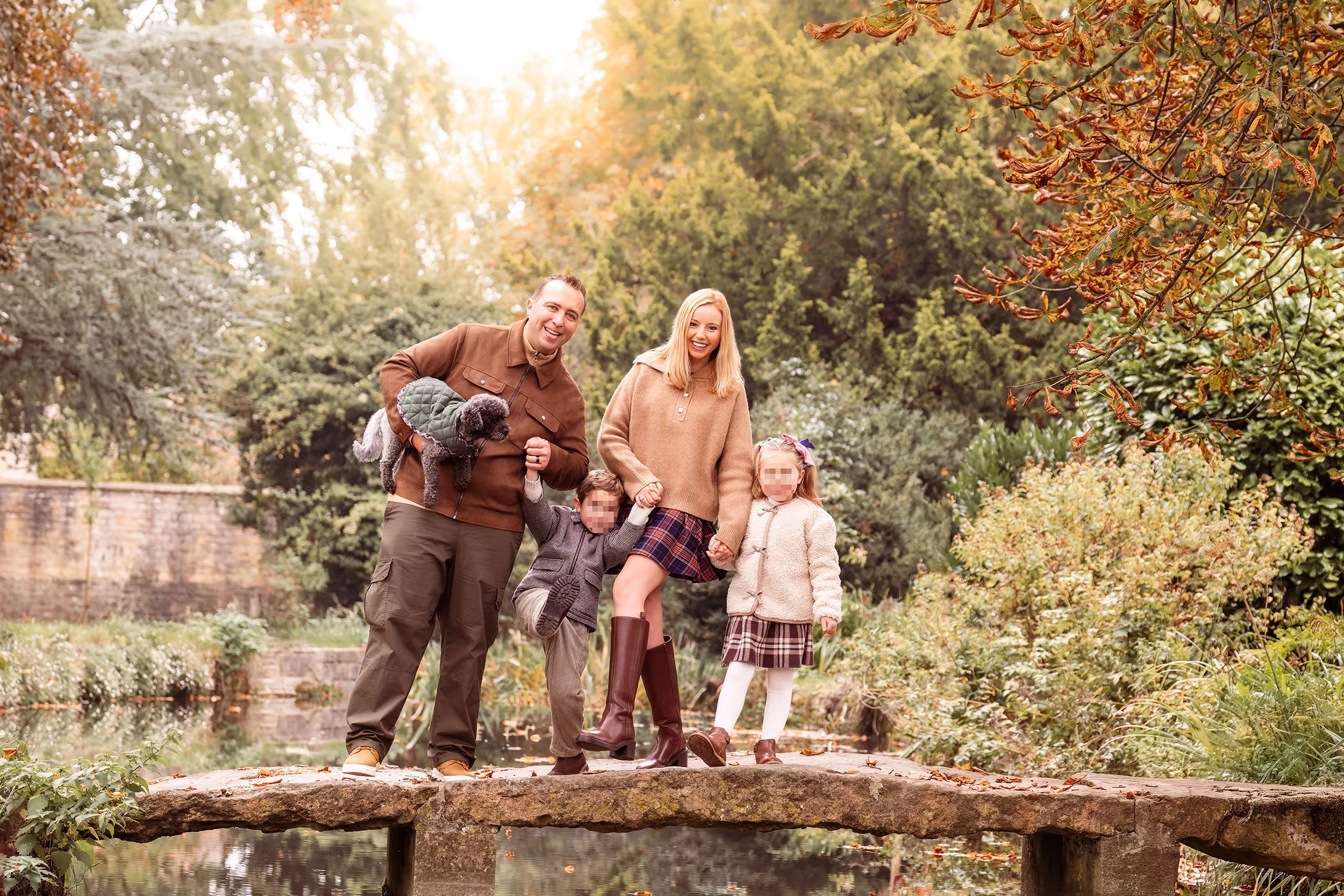 A family of four in a relaxed walking pose on a stone bridge, with warm glowing backlight, in a cute Cotswold village near Bourton on The Water.