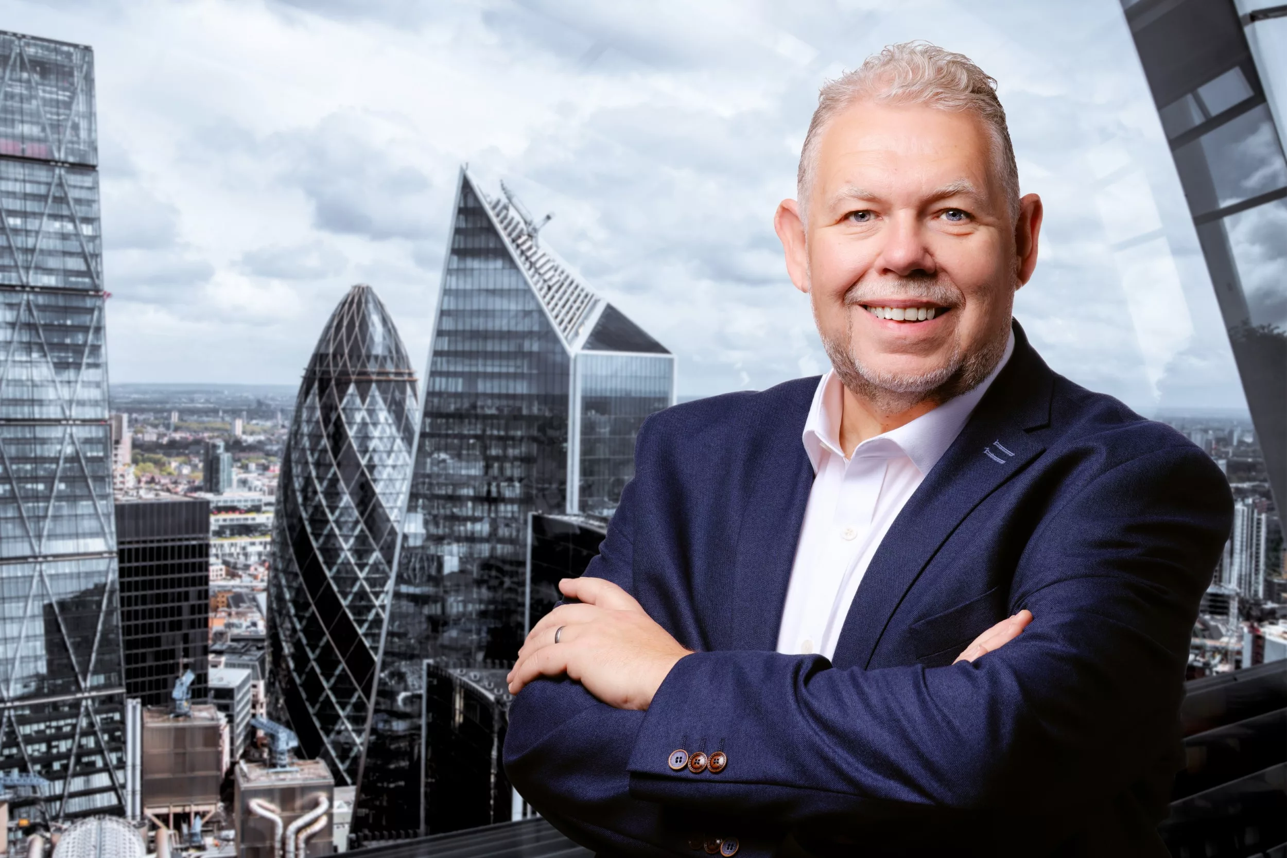A polished-looking businessman in a suit with his arms crossed, posing for his executive portrait, with the City of London skyline in the background.