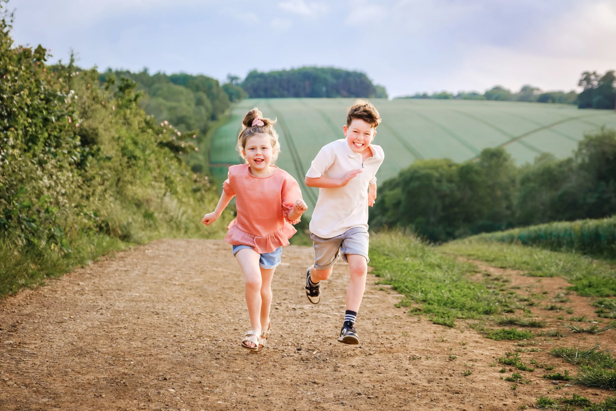 Two happy children running towards the camera, racing each other, with a beautiful hilly countryside background and soft light during sunset.