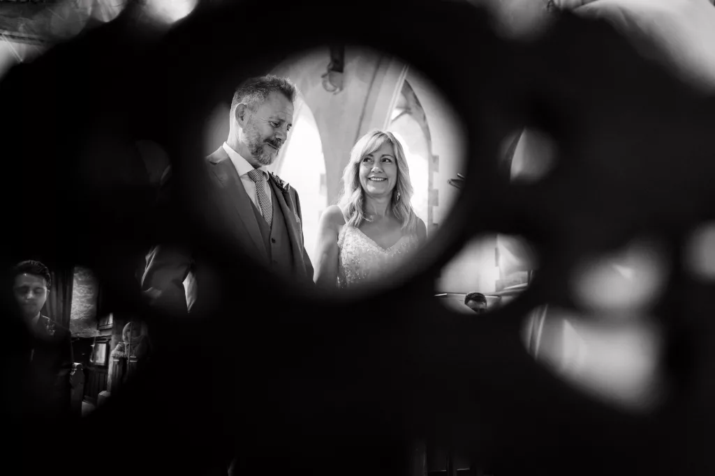 A bride and groom sharing a cheeky look during the ceremony, seen through a circular hole in a chair in the foreground.