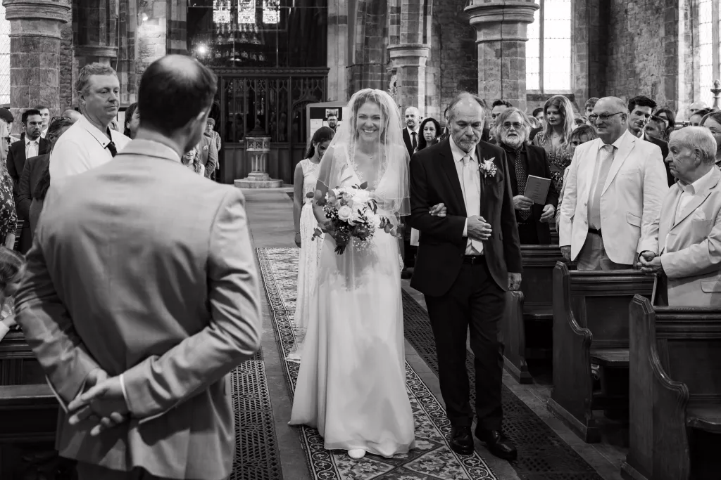 Bride grinning at groom while walking down the aisle with her father